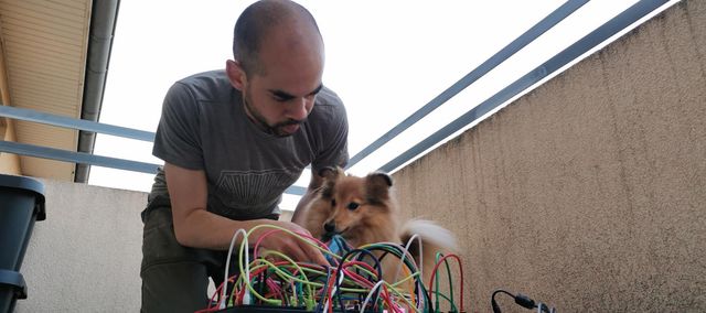 Photo d'un homme au dessus d'un synthé modulaire qui semble montrer à son chien ce qu'il fait